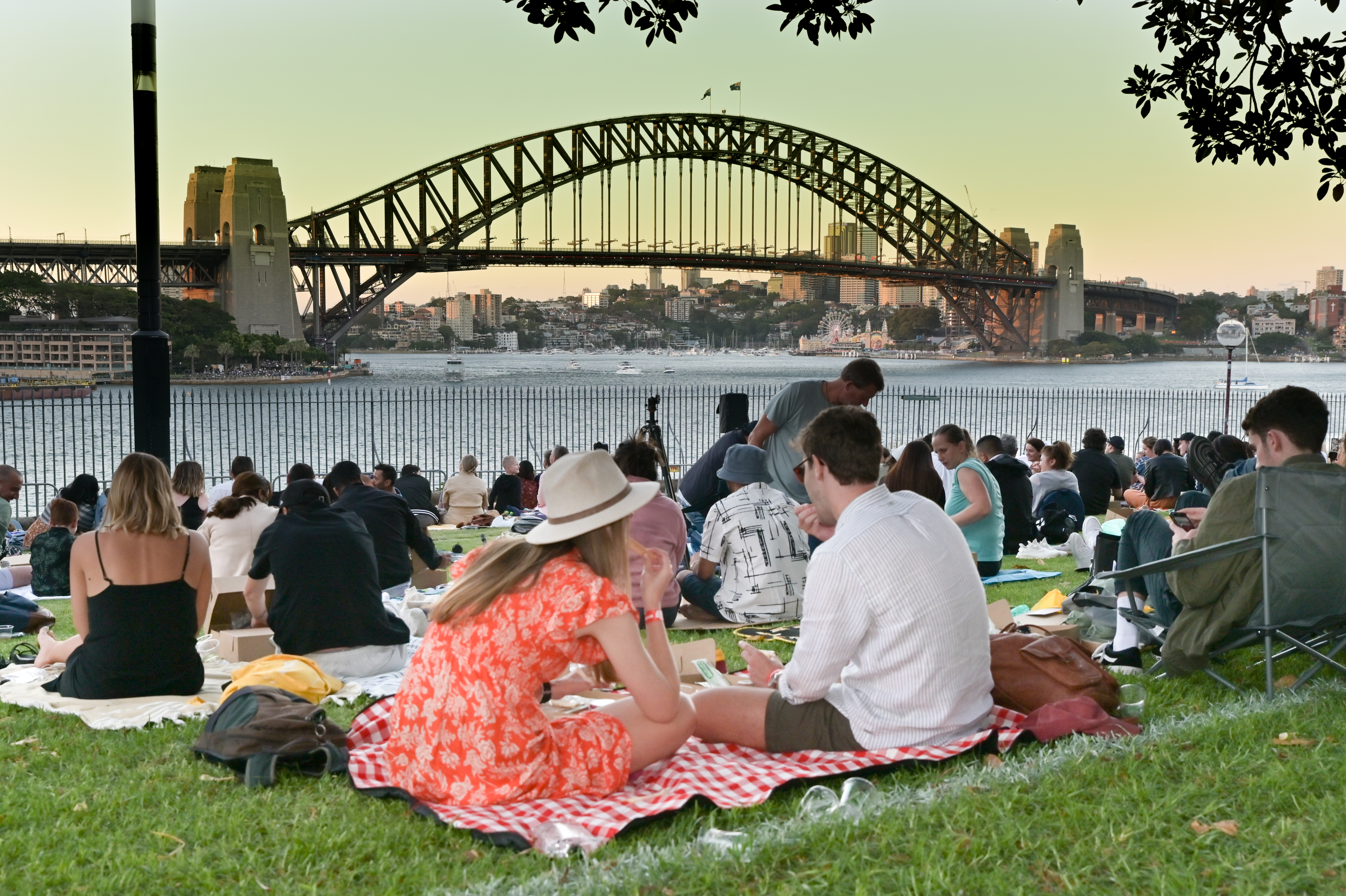 People sit on blankets having a picnic in a park near the water, with the Sydney Harbour Bridge in the background at sunset.