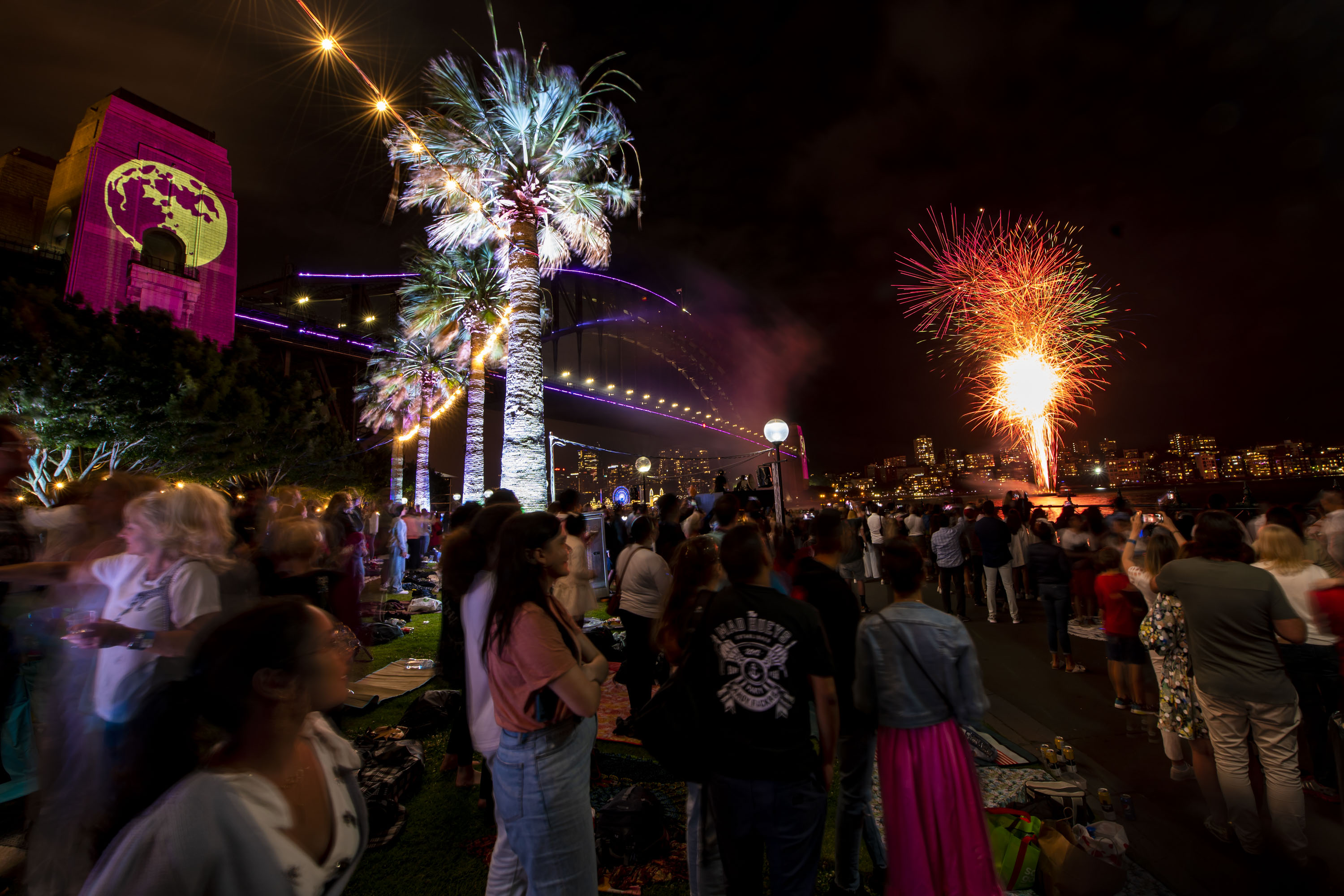 A crowd watches fireworks over the water near a lit-up bridge at night, with palm trees and colourful projections visible in the scene.