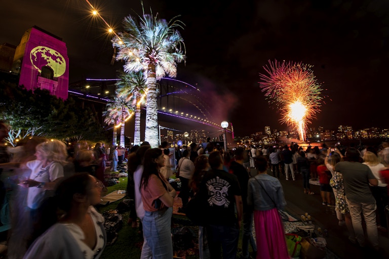 A crowd watches fireworks over the water near a lit-up bridge at night, with palm trees and colourful projections visible in the scene.