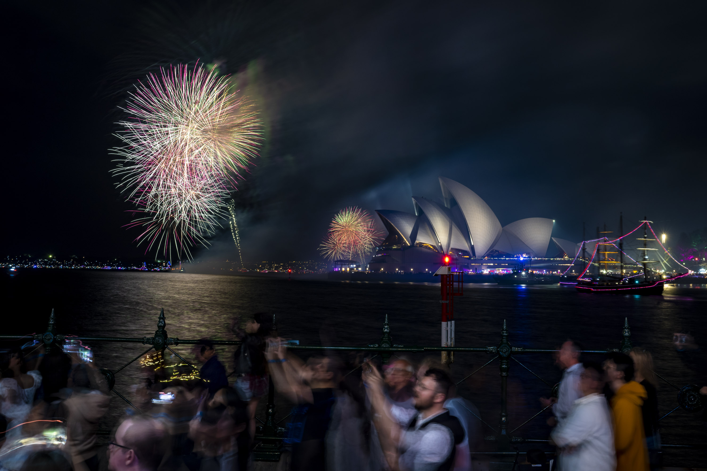Crowd watches colourful fireworks over Sydney Harbour, with the Sydney Opera House and a lit-up ship visible in the background at night.