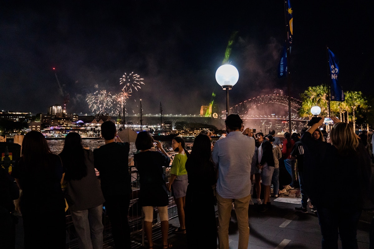 A crowd watches fireworks over Sydney Harbour at night, with the Sydney Harbour Bridge and city skyline illuminated in the background.