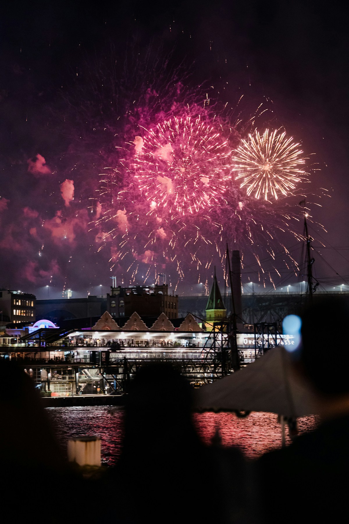 Colorful fireworks explode in the night sky over a waterfront cityscape, with silhouettes of people watching from the shore.