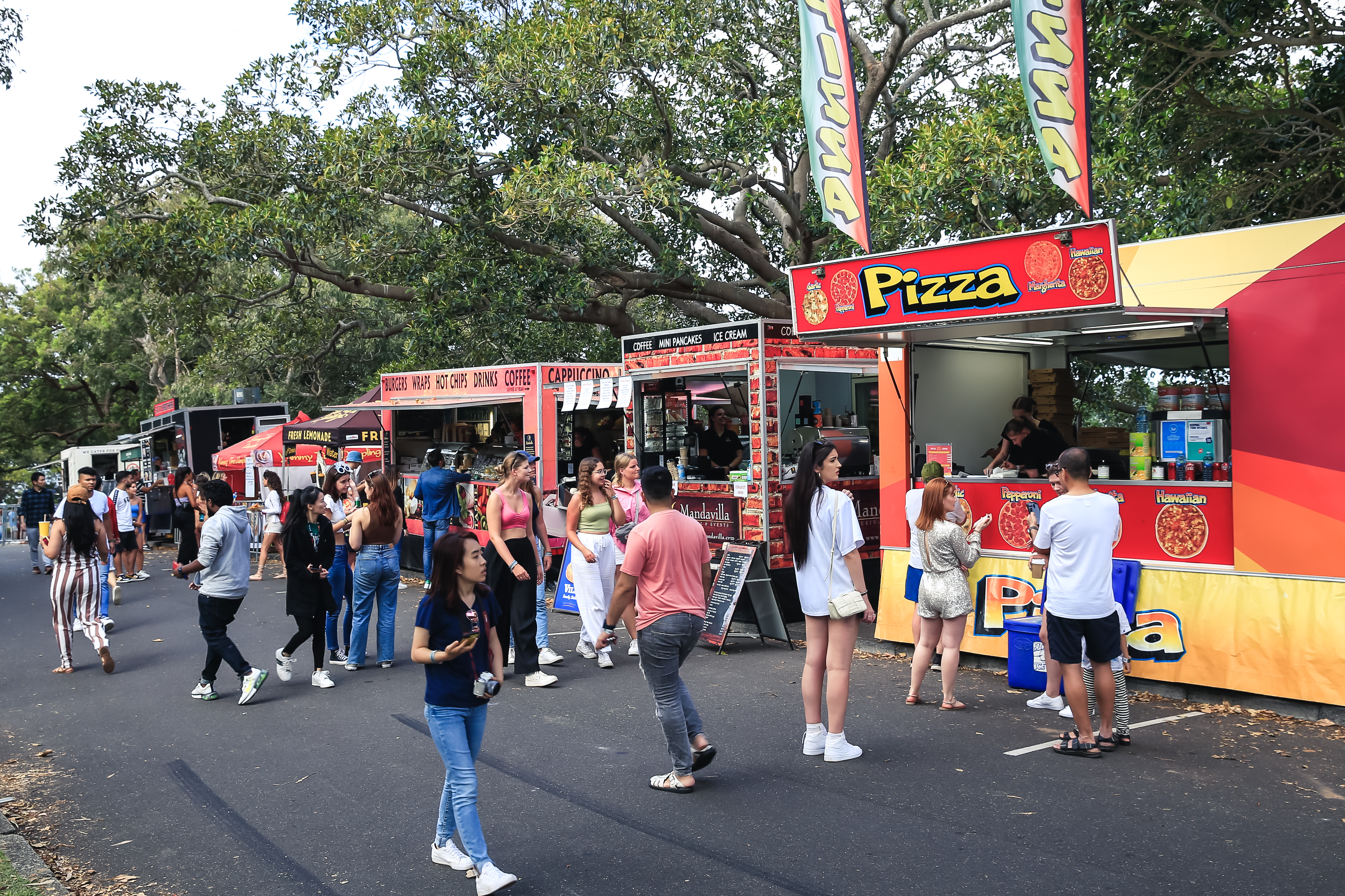 People gather at a food van event in a park, standing in queues and walking near various colourful food vans, including one selling pizza. Trees provide shade overhead.