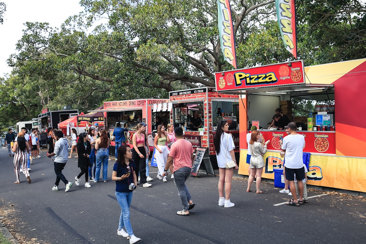 People gather at a row of colorful food trucks in a park, with signs advertising pizza and other foods. Trees and crowds fill the background.