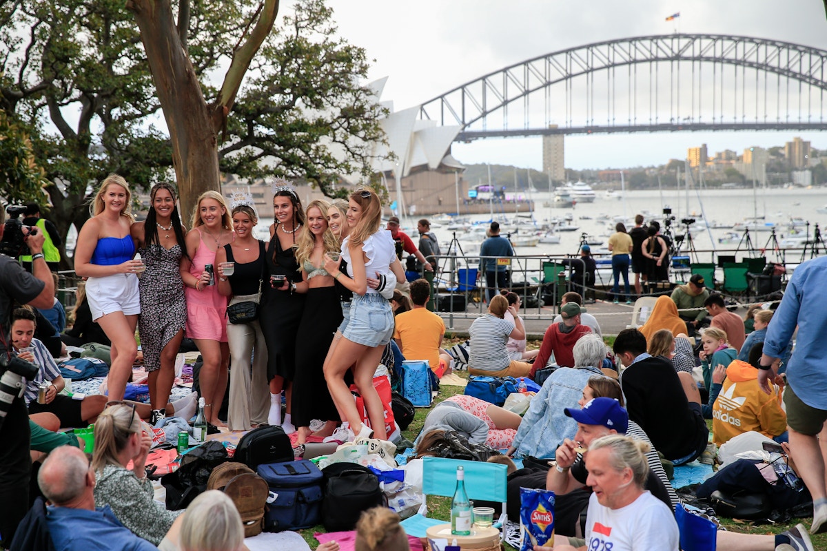A group of people picnic outdoors with the Sydney Opera House and Harbour Bridge in the background. Some pose for a photo while others sit on blankets.