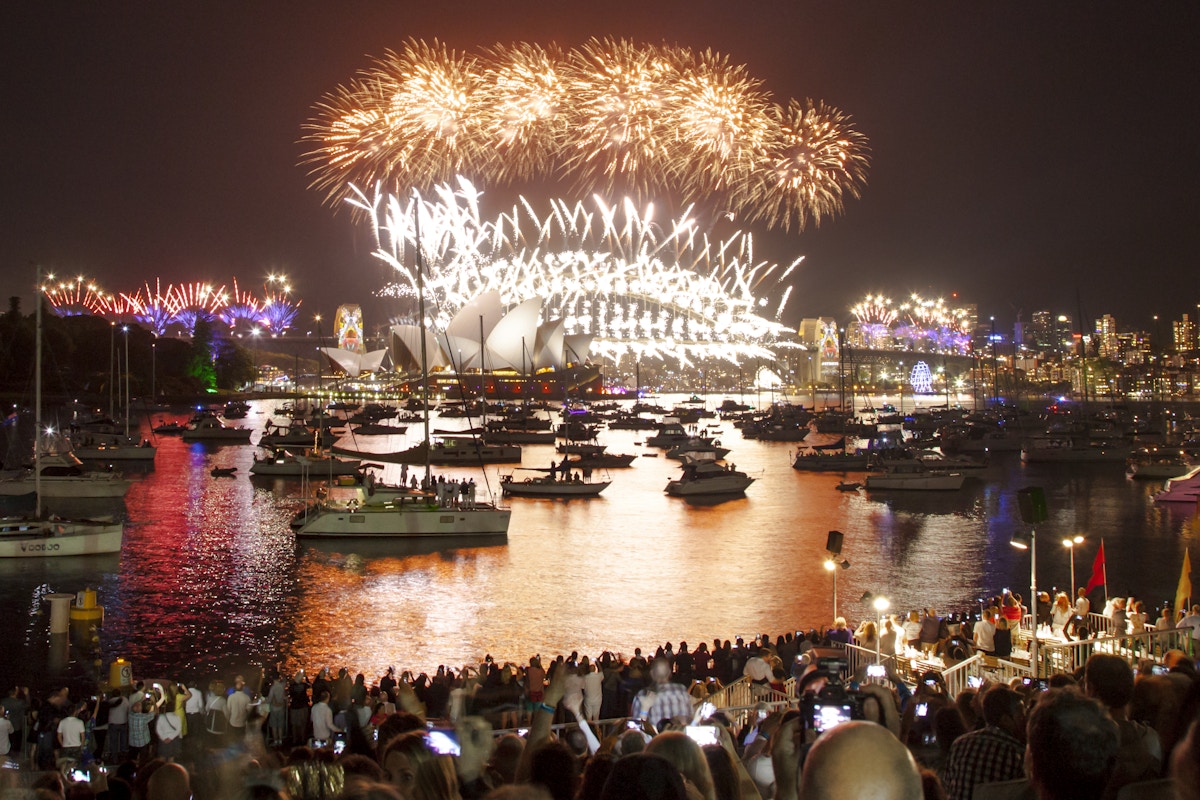 Crowds watch fireworks illuminating the night sky above Sydney Opera House and harbor, with boats on the water and city lights in the background.