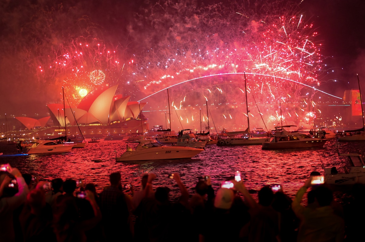 Fireworks light up the night sky over Sydney Opera House and Harbour Bridge, with boats on the water and crowds watching from the shore.