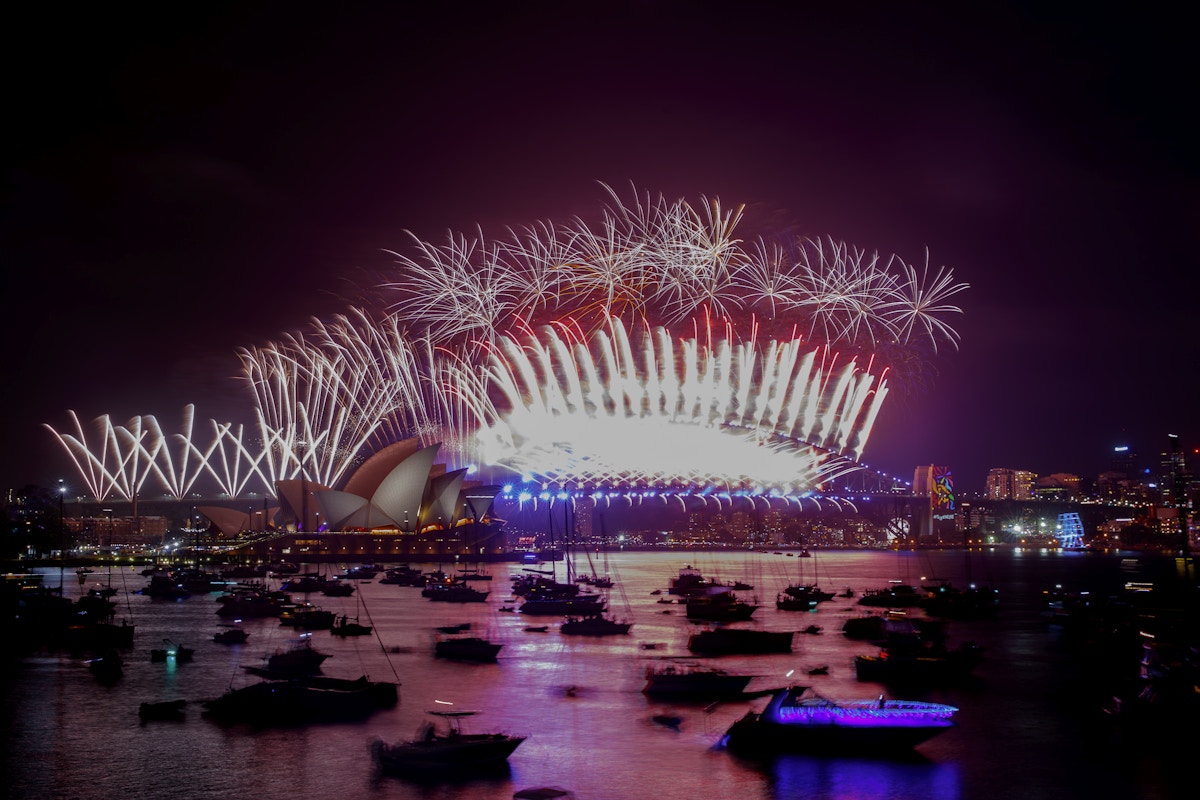Fireworks light up the night sky above Sydney Opera House and Harbour Bridge, with boats gathered on the water in the foreground.