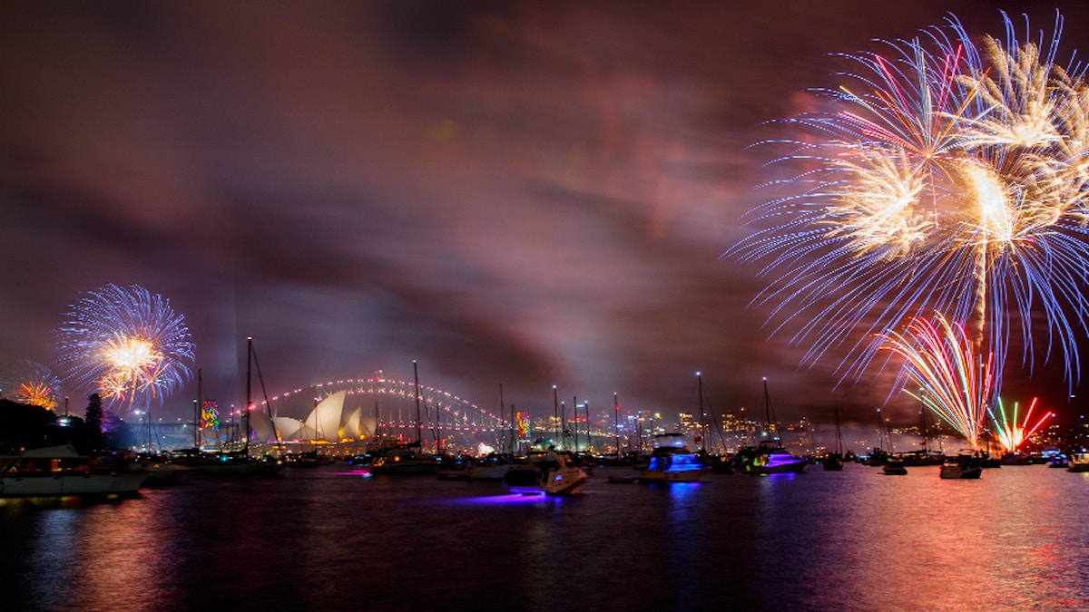 Fireworks light up the night sky over Sydney Harbour, with the Opera House, Harbour Bridge, and boats visible on the water.
