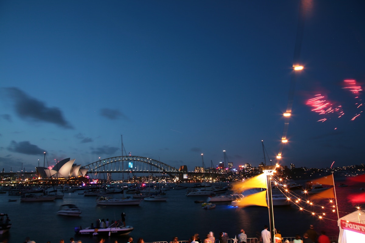 Boats gather on the water at dusk near Sydney Opera House and Harbour Bridge, with city lights and fireworks in the sky.