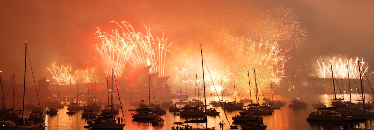 Fireworks light up the sky above Sydney Opera House as boats gather in the harbor at night, reflecting the bright colors on the water.