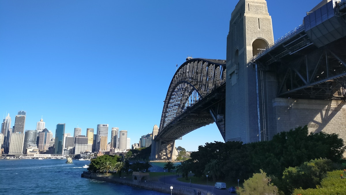 Sydney Harbour Bridge with city skyline in the background, blue sky, and water in the foreground.