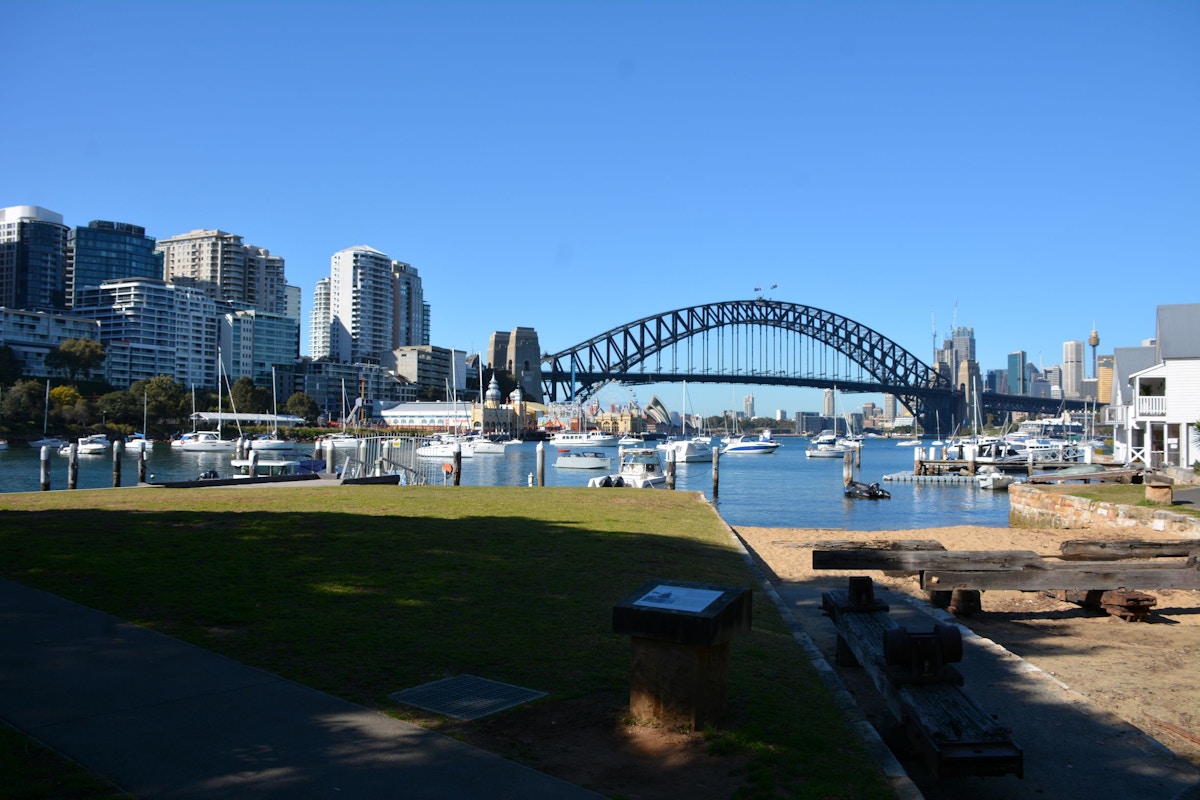 Sydney Harbour Bridge over the water with boats docked nearby, modern buildings on the left, and clear blue sky in the background.