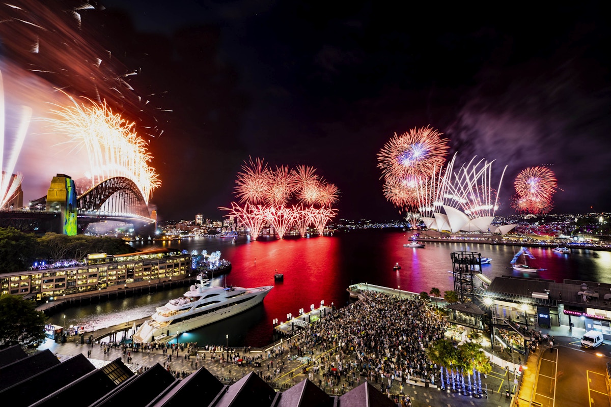 Fireworks light up the night sky over Sydney Harbour, with the Sydney Opera House and Harbour Bridge visible, and crowds gathered along the waterfront.