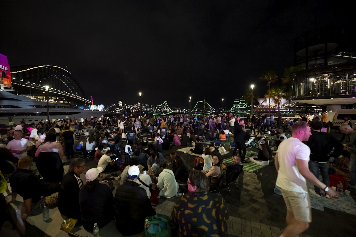 A large crowd gathers on a waterfront promenade at night, with boats decorated in lights and a lit-up bridge visible in the background.