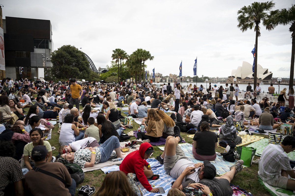 A large crowd of people sits and lounges on blankets at a grassy park beside the water, with the Sydney Opera House visible in the background.
