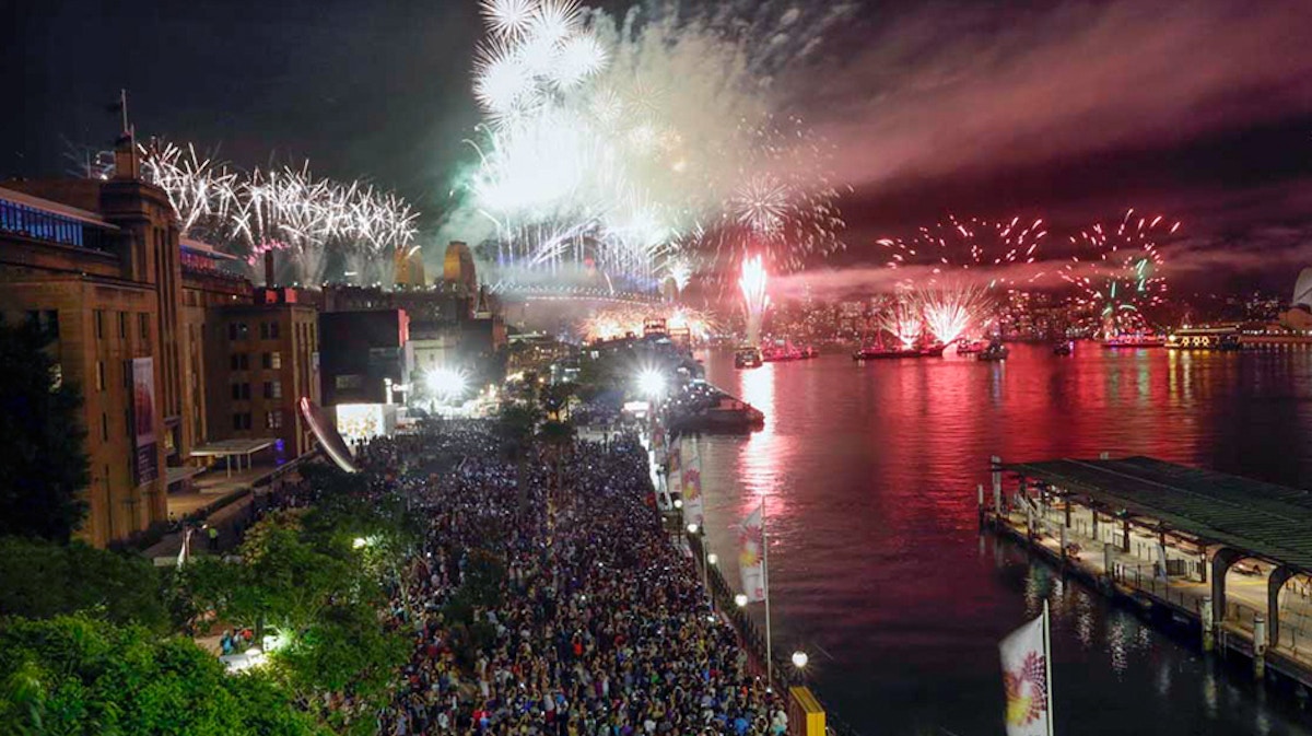 Large crowd gathers by the waterfront at night, watching colorful fireworks explode over a bridge and harbor.