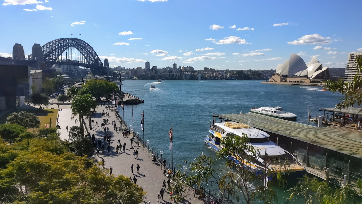 Sydney Harbour view with the Harbour Bridge on the left, the Opera House on the right, boats in the water, and people walking along the waterfront.