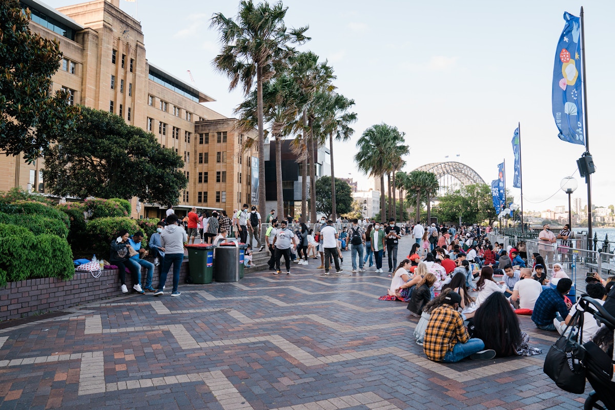 Groups of people sit and stand along a riverside promenade with palm trees, banners, and a view of a large bridge in the background.