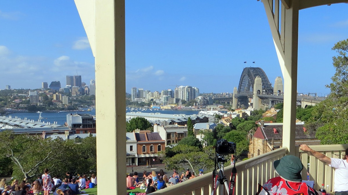 People sit on a grassy hill overlooking Sydney Harbour and the Sydney Harbour Bridge on a sunny day, with city buildings visible in the background.