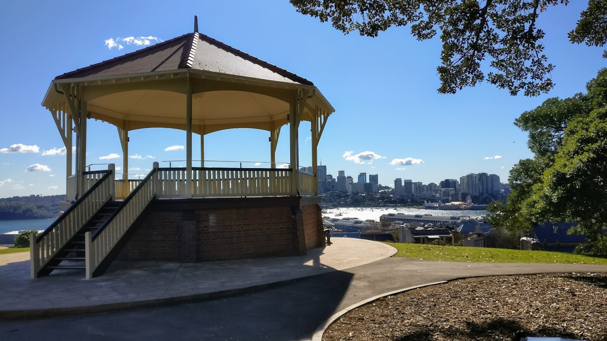 A gazebo stands on a hill in a park, with a view of a city skyline and water in the background under a clear blue sky.