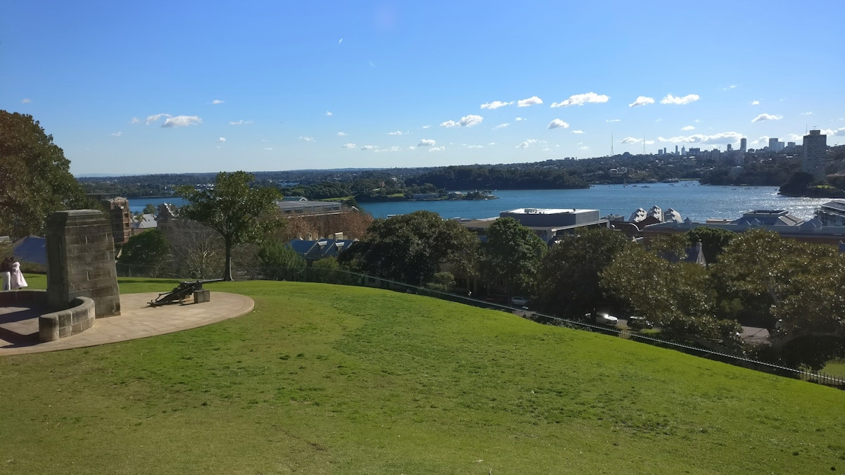 A grassy hill with a stone monument overlooks a river, trees, and city skyline under a clear blue sky with scattered clouds.