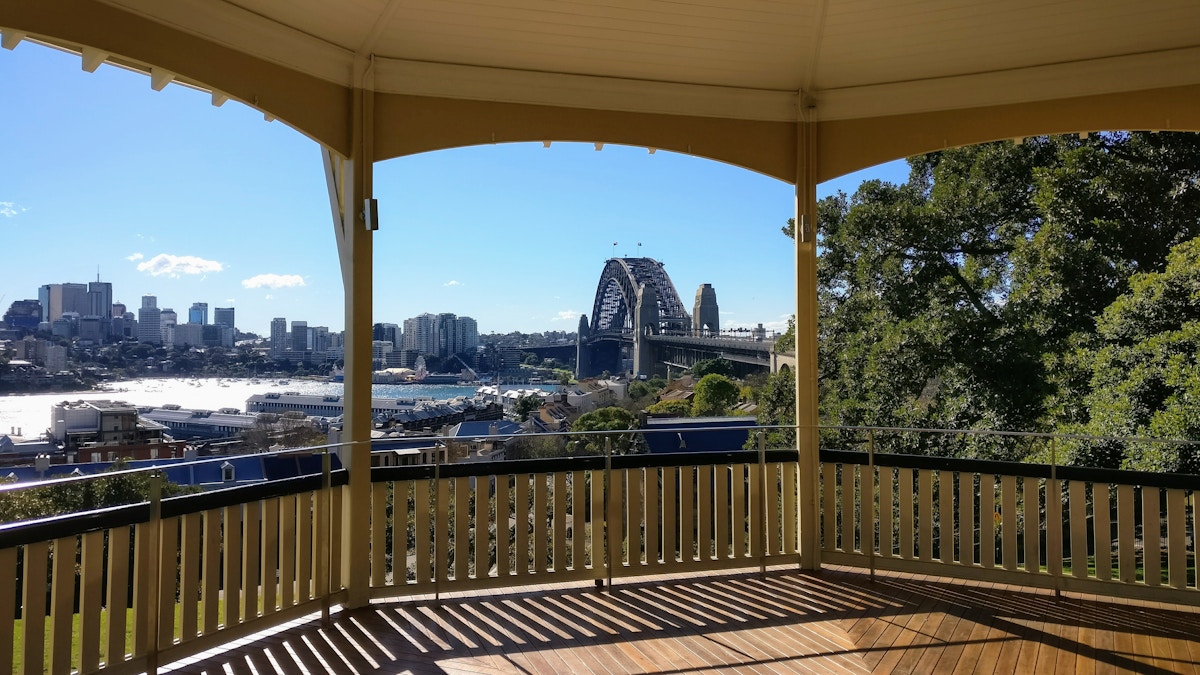 View of Sydney Harbour Bridge and city skyline seen from inside a gazebo, with wooden floor and railing in the foreground and trees partially visible on the right side.