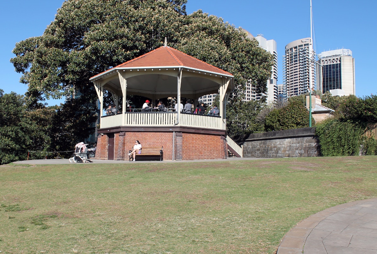 A brick and wood gazebo with a red roof sits on a grassy area, with people inside. City buildings and large trees are in the background under a clear blue sky.
