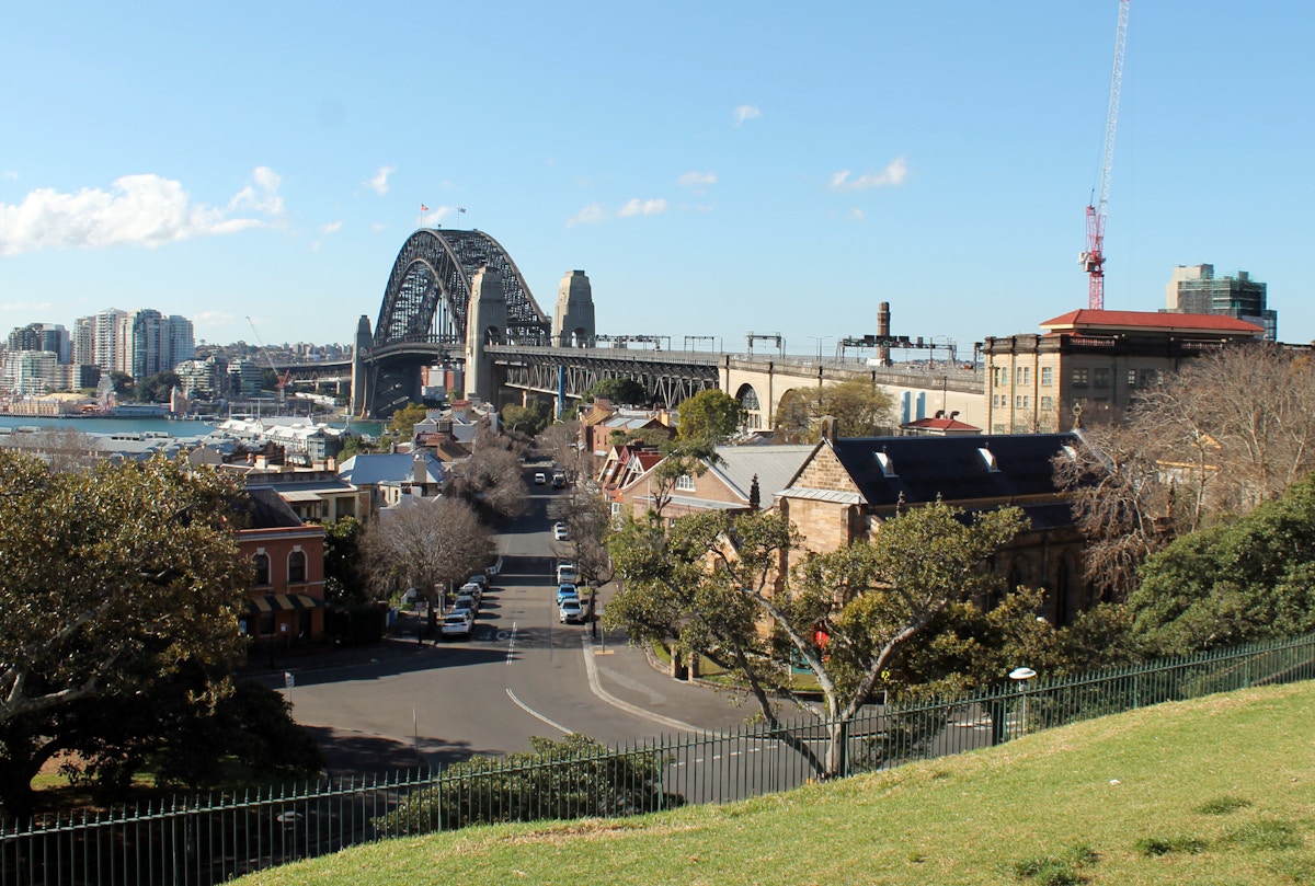A view of Sydney Harbour Bridge with nearby buildings, trees, and a street in the foreground under a clear sky.