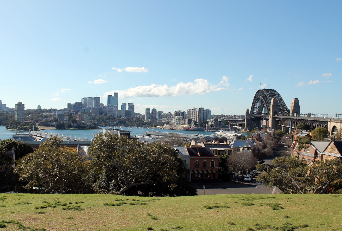 A view of Sydney Harbour with the Sydney Harbour Bridge on the right, city skyline in the background, and trees and buildings in the foreground under a clear sky.