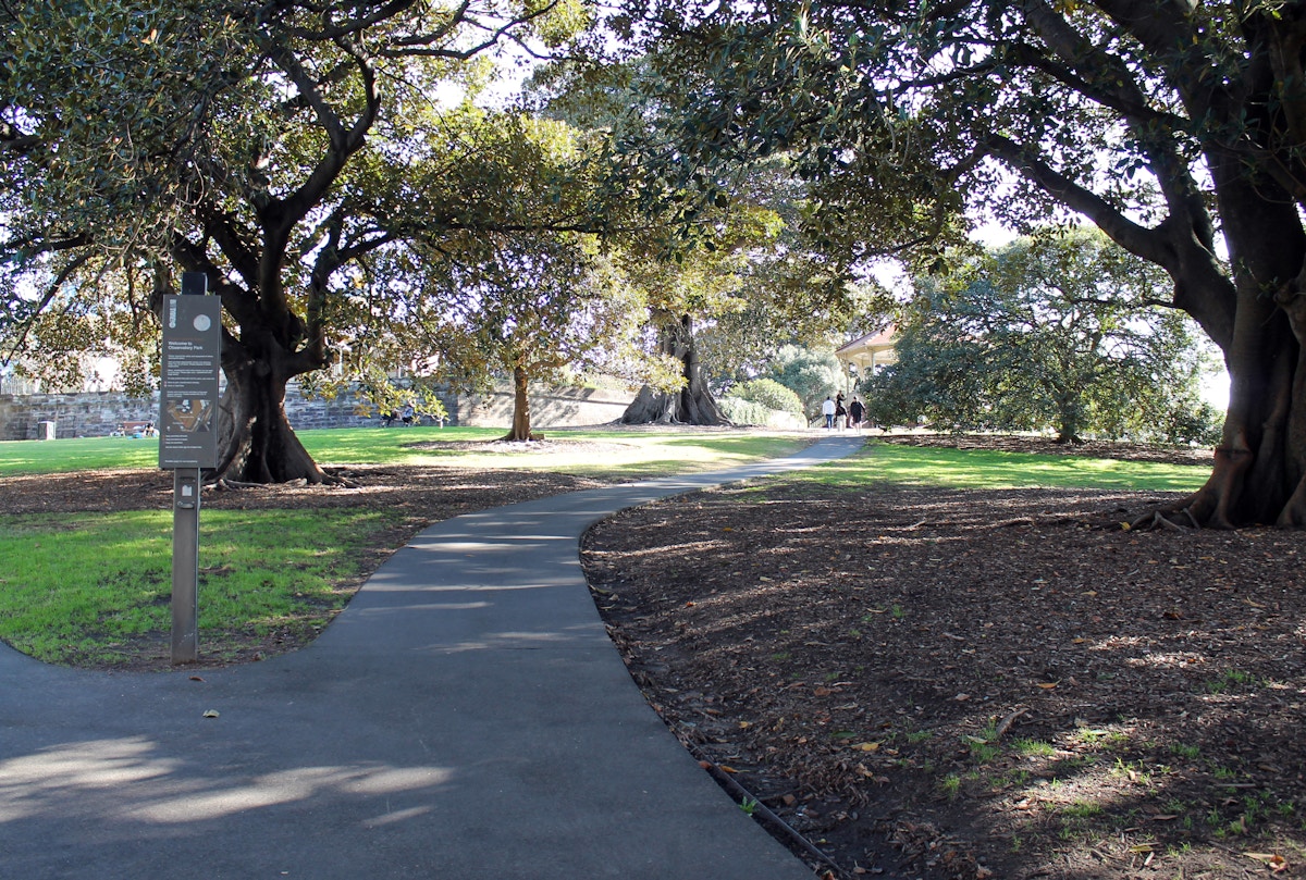 Paved path curves through a park with large trees, green grass, a signpost on the left, and a few people walking in the distance.