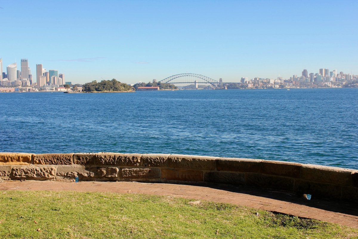 A view of Sydney Harbour with the Sydney Harbour Bridge and city skyline in the background, seen from a grassy area behind a stone wall.