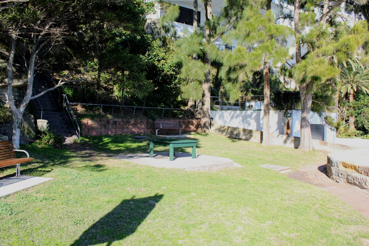 A small park area with a ping pong table, benches, trees, grass, and a staircase leading up a hillside in the background.