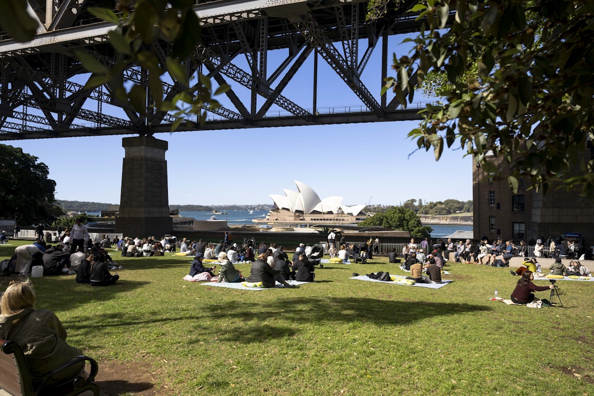 People sit on the grass under Sydney Harbour Bridge, with the Sydney Opera House and harbor visible in the background on a sunny day.