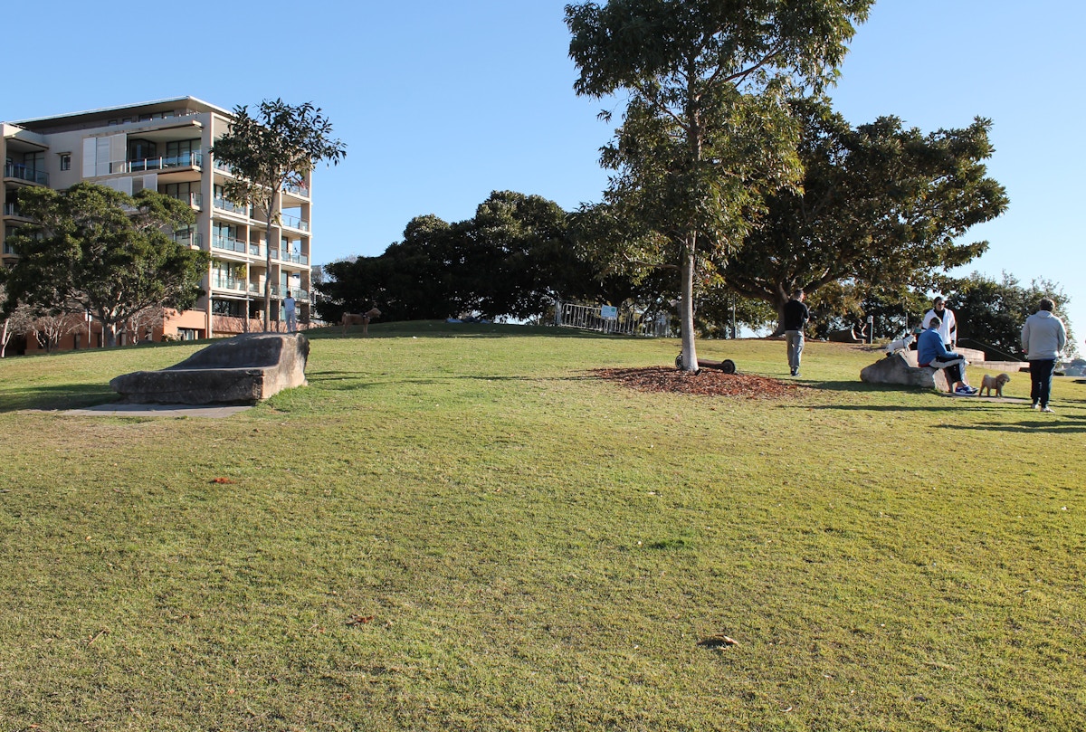 A grassy park with trees, large rocks, and several people walking or sitting. An apartment building is visible on the left under a clear sky.
