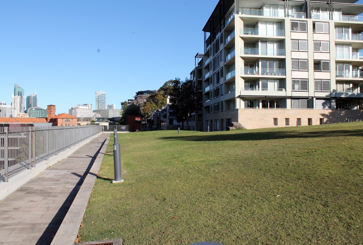 A modern apartment building stands beside a large grassy area, with a paved walkway and city skyscrapers visible in the distant background under a clear blue sky.