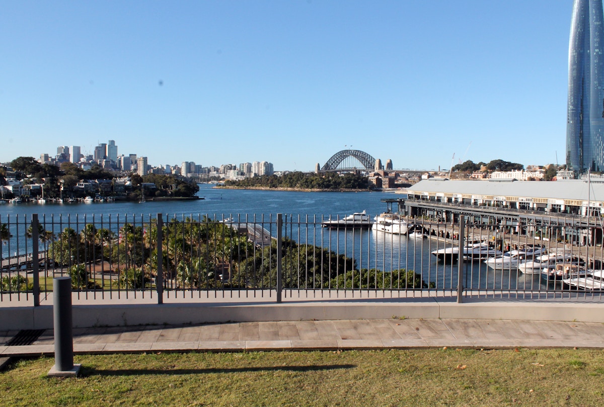 A waterfront view with a metal fence in the foreground, boats docked at a marina, and a city skyline with a large arched bridge in the background under a clear blue sky.