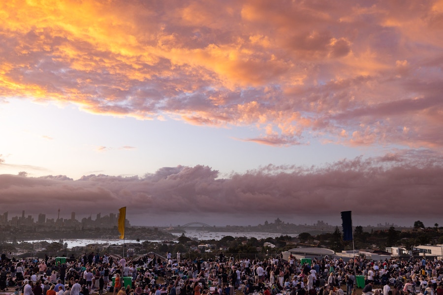 Large crowd gathers outdoors at sunset, with dramatic orange clouds in the sky and a distant city skyline visible on the horizon.