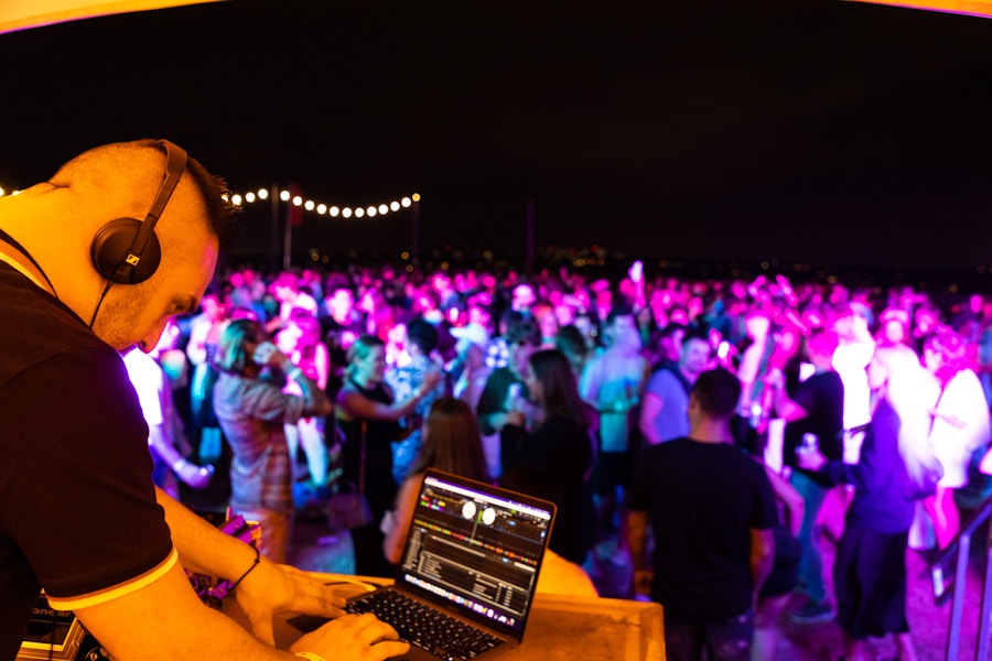 A DJ wearing headphones mixes music on a laptop while a large crowd dances under colorful lights at an outdoor nighttime event.