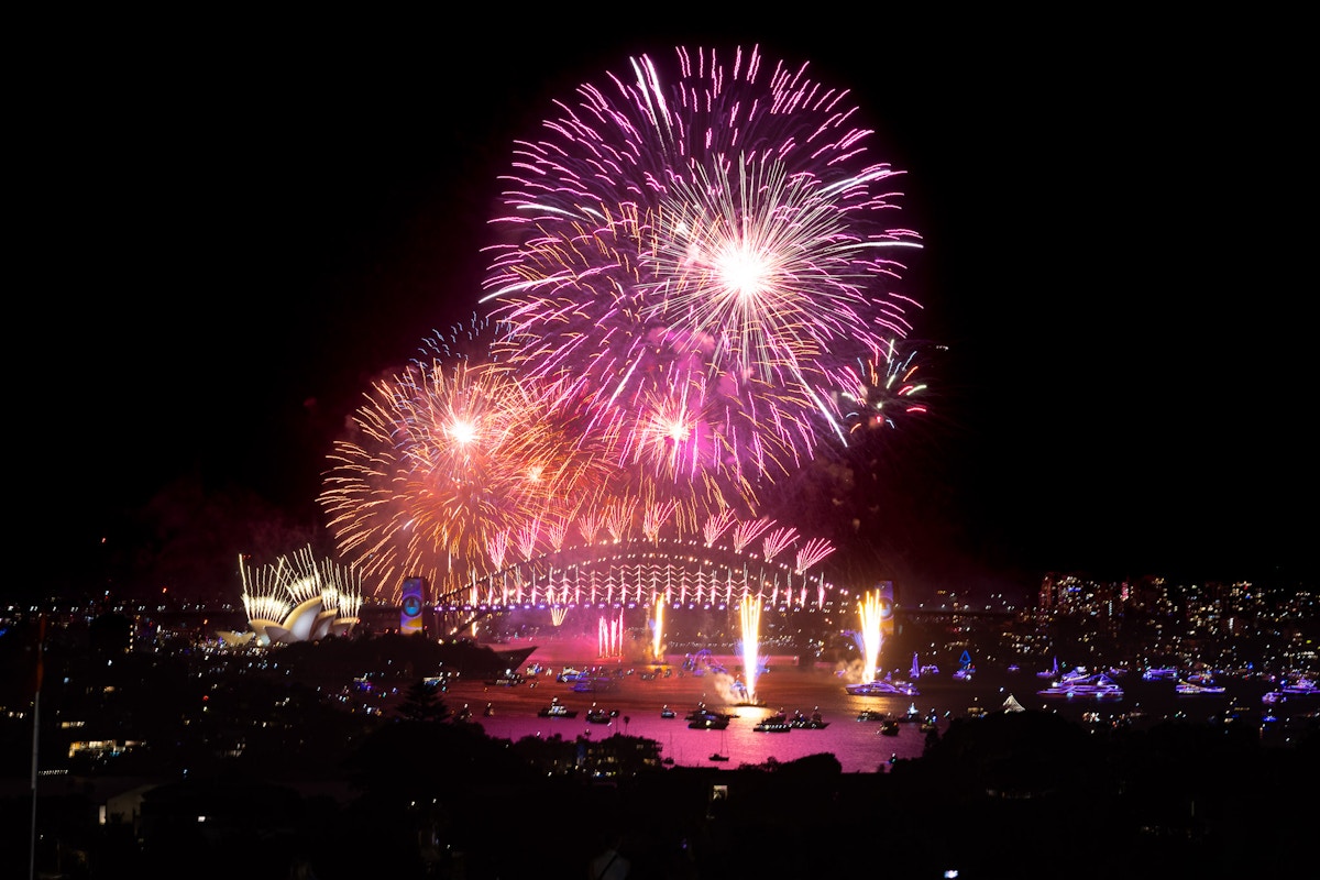 Fireworks display over Sydney Harbour at night, with the Sydney Opera House and Harbour Bridge illuminated, and boats visible on the water.
