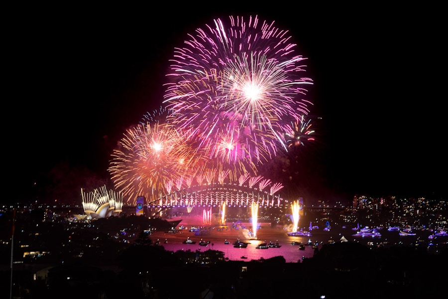 Fireworks display over Sydney Harbour at night, with the Sydney Opera House and Harbour Bridge illuminated, and boats visible on the water.