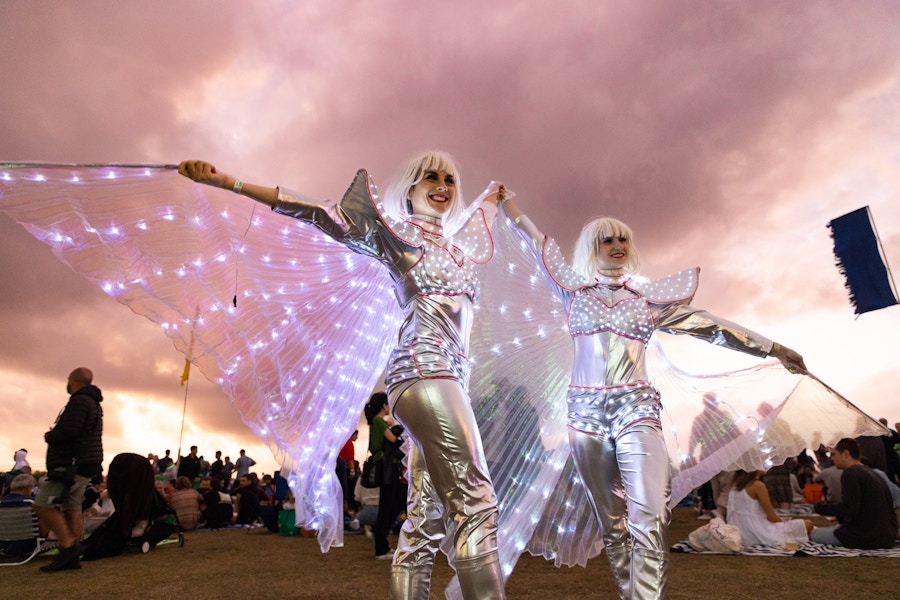 Two people in silver costumes with illuminated wings pose outdoors at an event, with a cloudy sky and a crowd in the background.
