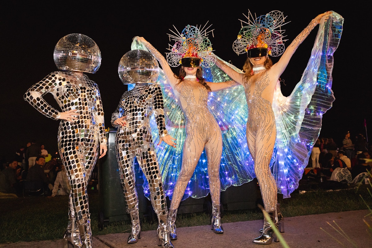 Four people in reflective costumes and headpieces pose at night; two have disco ball helmets, two wear illuminated winged outfits and colorful spiked hats.