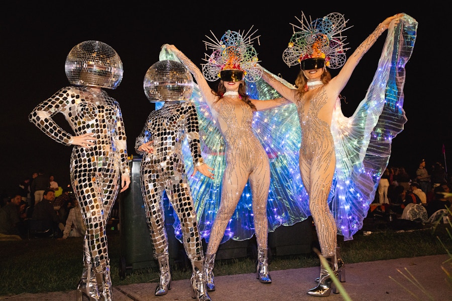 Four people in reflective costumes and headpieces pose at night; two have disco ball helmets, two wear illuminated winged outfits and colorful spiked hats.