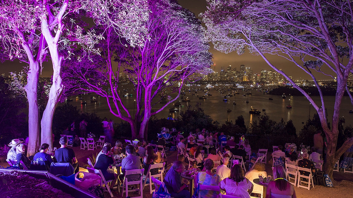 People sit at tables outdoors under trees lit with purple lights, overlooking a harbor with boats and a city skyline in the background at night.