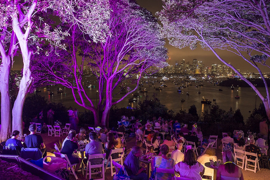 People sit at tables outdoors under trees lit with purple lights, overlooking a harbor with boats and a city skyline in the background at night.
