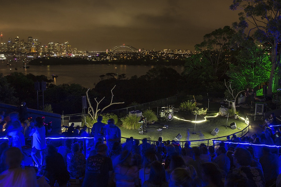 An outdoor stage set with musical instruments overlooks Sydney Harbour at night, with an audience seated and city lights in the background.