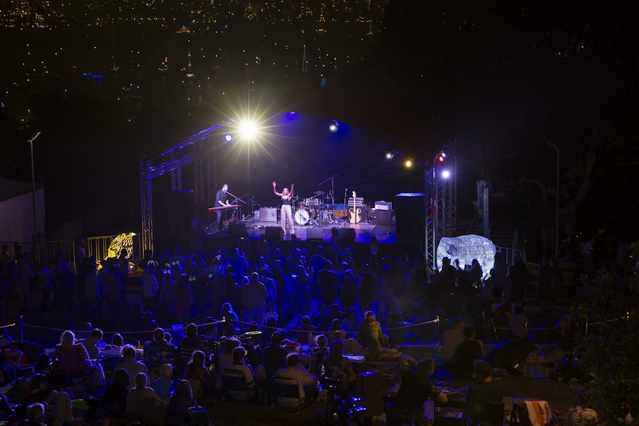 A band performs on a lit outdoor stage at night, facing an audience seated on the grass and standing in front of the stage, with city lights visible in the background.