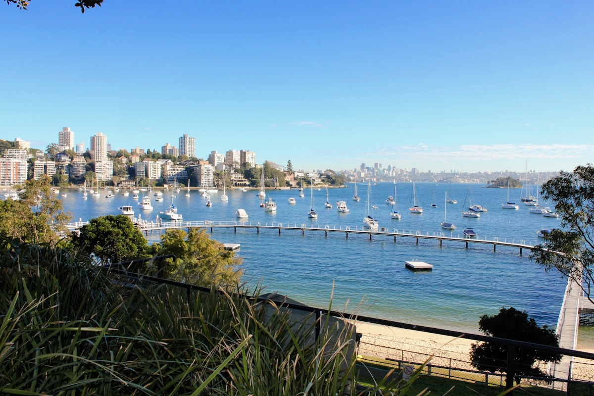 Sailboats anchored in a calm bay with a pier, sandy beach, high-rise buildings, and city skyline in the background under a clear blue sky.