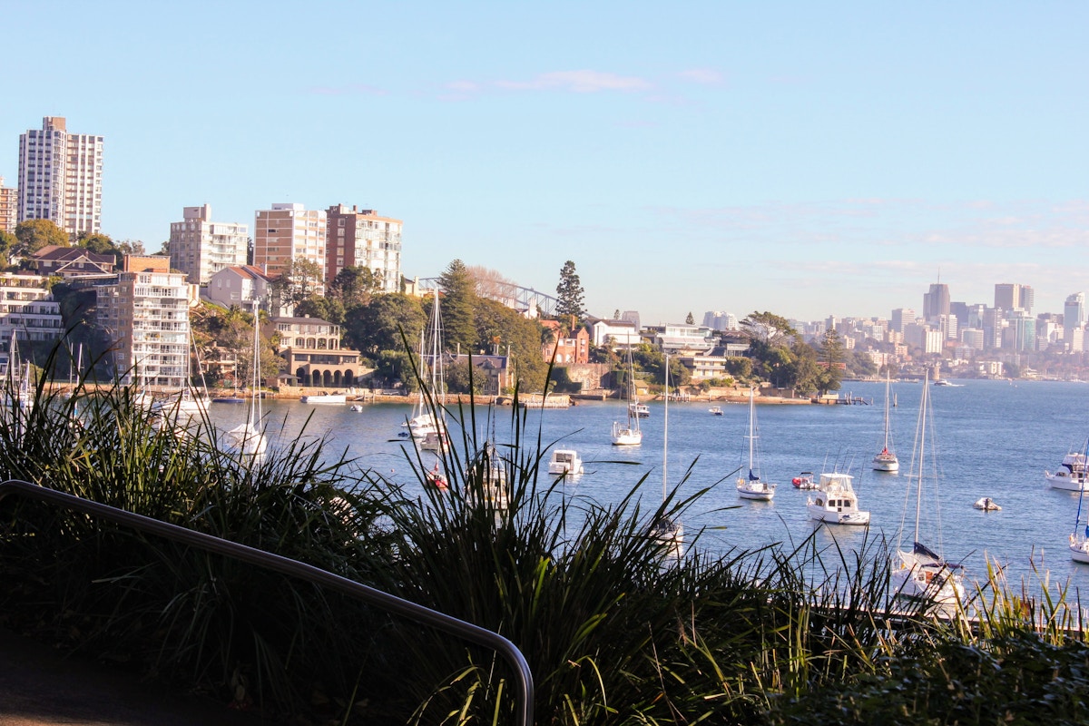 View of a harbor with moored boats, city buildings, and greenery along the shoreline under a clear blue sky.